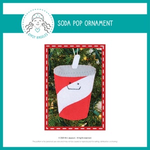 May include: A felt Christmas ornament shaped like a soda pop cup with a red and white striped design. The cup has a straw and a smiling face. The ornament is hanging on a green Christmas tree.
