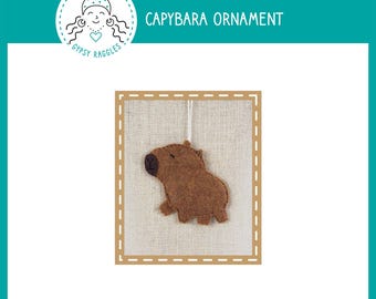May include: A brown felt capybara ornament suspended by a white string, presented on a beige fabric backdrop within a brown frame. The text "CAPYBARA ORNAMENT" is above the ornament. The Gypsy Raggles logo is in the upper left corner.