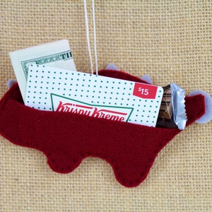 May include: Red felt dinosaur ornament with a Krispy Kreme gift card, a chocolate bar, and cash inside. The ornament has a loop for hanging.