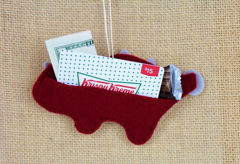 May include: Red felt dinosaur ornament with a Krispy Kreme gift card, a chocolate bar, and cash inside. The ornament has a loop for hanging.