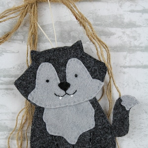 May include: A gray felt wolf ornament with a white string for hanging. The wolf has a stitched smile and white teeth.