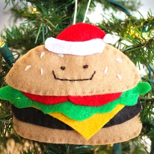 May include: A felt Christmas ornament shaped like a hamburger with a red and white Santa hat. The hamburger has a smiling face and is decorated with green lettuce, red tomato, yellow cheese, and a black patty.
