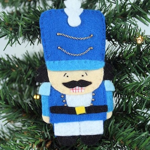 May include: A felt Christmas ornament shaped like a toy soldier. The ornament is primarily blue with gold chain accents, a black mustache, and a white plume. The soldier has a black belt with a yellow buckle and is hanging on a Christmas tree.