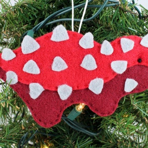 May include: Red and burgundy felt Christmas ornament shaped like an ankylosaurus dinosaur with white felt spikes and a black eye.