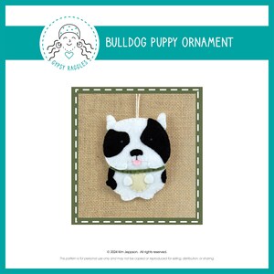May include: A felt bulldog puppy ornament with black and white fur, a pink tongue, and a green collar. The ornament is hanging from a string and is on a burlap background.