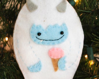 Puede incluir: Adorno de fieltro blanco con forma de yeti sosteniendo un cono de helado rosa. El yeti tiene ojos azules y una sonrisa cosida.