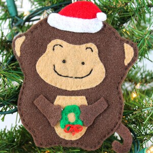 Puede incluir: Un adorno de mono de fieltro marrón que lleva un gorro de Papá Noel rojo y blanco y sostiene una corona de fieltro verde. El adorno está colgado de un árbol de Navidad verde.