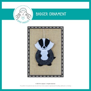 May include: A felt badger ornament with a black, white, and gray design. The badger has a white face and a black stripe down its back. The ornament hangs from a white string and is displayed on a burlap background. The text "BADGER ORNAMENT" is above the ornament.