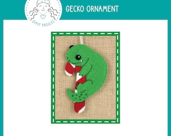 May include: A green felt gecko ornament holding a red and white candy cane. The ornament has a loop for hanging and is labeled "Gecko Ornament".
