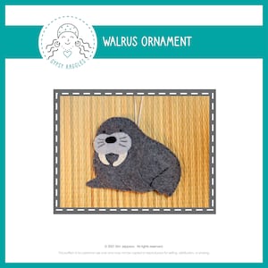 Puede incluir: Un adorno de foca barbuda de fieltro gris con colmillos blancos y una nariz gris. El adorno está colgado de una cuerda blanca y se encuentra sobre un fondo de madera marrón. El texto "WALRUS ORNAMENT" está en la parte superior de la imagen.