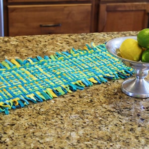 May include: A woven table runner with blue, green, and yellow stripes. The runner has a fringe edge and is displayed on a granite countertop with a silver bowl of lemons and limes.