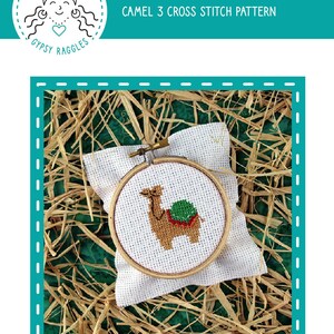 May include: A cross-stitch pattern featuring a camel design, displayed in a wooden embroidery hoop. The camel is brown with a green saddle and red accents. The text "CAMEL 3 CROSS STITCH PATTERN" is visible. The background is straw.