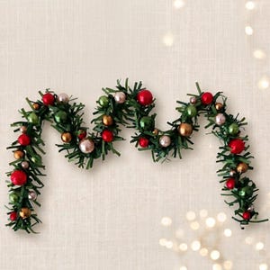 May include: A festive garland featuring dark green artificial pine needles interspersed with small, round ornaments in shades of red, metallic gold, olive green, and pale pink, arranged in a wavy, draped pattern across the horizontal frame.