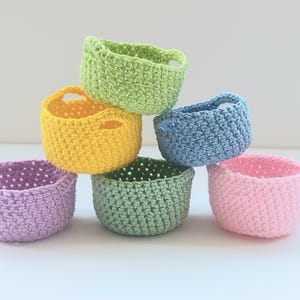 May include: A stack of six small, handmade crochet baskets in various colors: green, yellow, blue, pink, purple, and green. Each basket has a rounded shape with a textured stitch pattern and small handles.