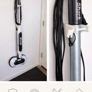 Puede incluir: Una mopa de vapor Shark blanca y plateada está montada en una pared blanca. La mopa se asegura con un soporte de pared y tiene un cable de alimentación negro. La imagen también muestra una solución de almacenamiento de cables.