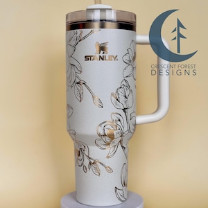 Peut inclure: Mug de voyage en acier inoxydable blanc avec un motif floral doré et une poignée blanche. Le mug porte le logo Stanley et le texte "Crescent Forest Designs".