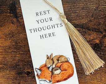 May include: A white rectangular bookmark featuring the black text REST YOUR THOUGHTS HERE and an illustration of a sleeping fox, a rabbit, and a hedgehog curled together. A gold-coloured tassel is attached through a hole at the top.