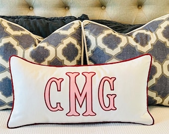 May include: Three decorative pillows on a bed. Two pillows have a geometric pattern in shades of gray and white. The third pillow is white with a red border and the monogram "CMG" embroidered in pink.