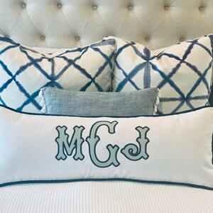 May include: A bed arrangement featuring decorative pillows. The primary pillow is white with a navy blue border and embroidered initials. Additional pillows include patterned and solid options in shades of blue and gray, creating a layered, stylish look.