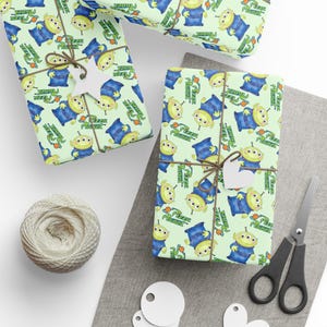 May include: Gift-wrapped presents with a light green background and a repeating pattern of blue aliens with three eyes. The gifts are tied with twine and adorned with white tags. A spool of twine and scissors are also visible.