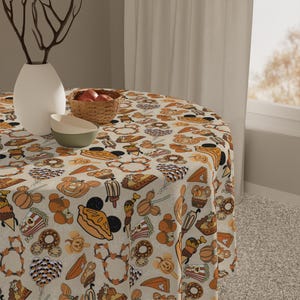May include: A round tablecloth featuring a repeating pattern of autumn-themed snacks and treats in shades of orange, brown, and cream. The design includes illustrations of pumpkin-shaped pastries, doughnuts, pies, popcorn, and balloons with mouse ears.