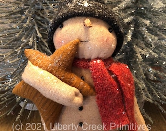 May include: A handmade snowman doll wearing a black hat and red scarf, holding a gold star. The doll is standing on a wooden surface.