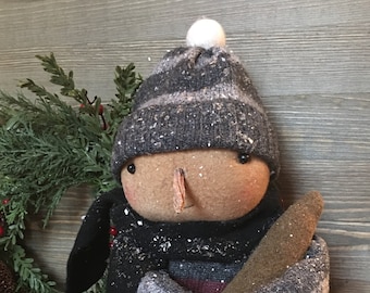 May include: A handmade rag doll snowman wearing a grey and white striped hat, a black scarf, and a grey sweater. The doll is holding a brown stick and is covered in white faux snow.