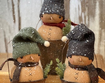 May include: Three handcrafted snowman figures in rustic metal pots. Each snowman has a fabric body, a stitched face, and wears a hat and scarf. The snowmen are decorated with faux snow and greenery, creating a charming winter scene.