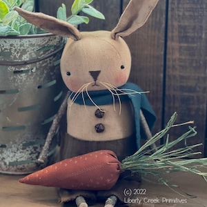 May include: A brown fabric bunny with a blue scarf and a carrot in its lap. The bunny has a brown nose and black eyes. The carrot is red and has green leaves. The bunny is sitting on a wooden surface. (c) 2022 Liberty Creek Primitives