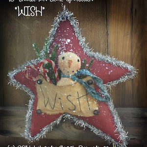 May include: A festive red star-shaped Christmas decoration featuring a snowman. It has silver tinsel edging and a burlap banner with the word "Wish." The snowman has a carrot nose and a blue scarf. The ornament is approximately 25 cm in diameter.