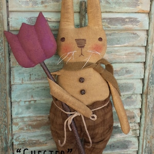 May include: A 16-inch brown bunny doll with a brown scarf and brown pants holding a purple tulip. The doll is standing in front of a wooden background. The text "CHESTER" and "16" BUNNY EPATTERN" is visible below the doll.