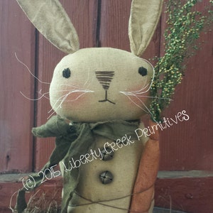 May include: A 15-inch beige rabbit doll with brown eyes, a green scarf, and a carrot. The doll is holding a carrot and has a bell on its chest. The doll is sitting in a nest of straw. The text "EDGAR" and "15" RABBIT PATTERN" is visible in the image. The text "(c) 2015 Liberty Creek Primitives" is also visible in the image.