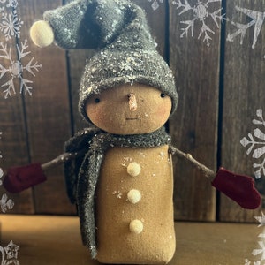 Puede incluir: Un muñeco de nieve hecho a mano que lleva un gorro y una bufanda de punto gris con guantes rojos. El muñeco tiene una cara marrón con botones blancos para los ojos y una nariz de pompón blanco. El muñeco está de pie sobre una superficie de madera con un patrón de copo de nieve blanco en el fondo. (2022) LIBERTY CREEK PRIMITIVES