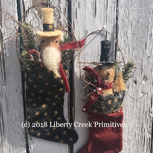 May include: Two Christmas stocking decorations. One features a Santa Claus figure with a white beard, red scarf, and a black stocking with gold stars. The other is a snowman with a black top hat and red scarf. Both are decorated with greenery.