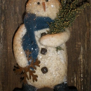 May include: A white snowman ornament with a blue scarf, holding a sprig of dried greenery. The snowman has a brown snowflake and bells on its body. The ornament is on a rustic wooden background.