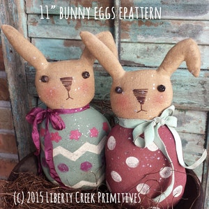 May include: Two 11 inch bunny egg patterns. One bunny is wearing a green and white egg pattern with a pink ribbon. The other bunny is wearing a red and white polka dot egg pattern with a light green ribbon. Both bunnies have brown eyes and a brown nose. The text "(C) 2015 Liberty Creek Primitives" is at the bottom of the image.