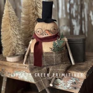 May include: A handmade snowman doll with a black top hat, round glasses, and a burgundy scarf. The snowman wears a plaid patterned outfit and has twig arms. The text "DIGITAL PATTERN" and "LIBERTY CREEK PRIMITIVES" are visible.