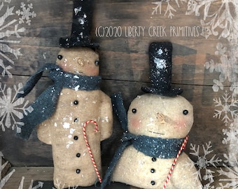 May include: Two handmade fabric snowmen with black top hats, grey scarves, and red and white candy canes. The snowmen are standing on a wooden surface with a white snowflake background.