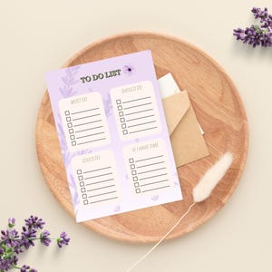 Op de afbeelding: Een lavendelkleurig 'To Do List' notitieblok met bloemmotieven, met secties voor taken. Het notitieblok ligt op een houten bord, met een kleine envelop en decoratieve lavendel takjes.