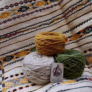 Peut inclure: Trois pelotes de laine, jaune moutarde, vert olive et blanc cassé, reposent sur un textile à motifs. Une petite étiquette avec les mots "FREE WILL" est visible. Le textile a un fond crème avec des motifs géométriques noirs, jaunes et rouges.