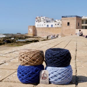 May include: Four skeins of yarn in shades of brown, navy blue, and two shades of blue are stacked on a stone surface. A small card with the text "FREE WILL" is placed in front of the yarn. The background shows a coastal scene.
