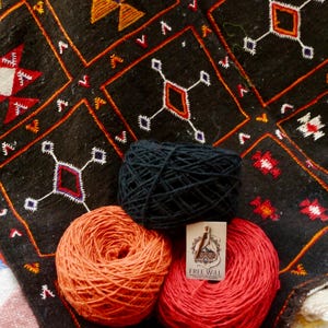 May include: Three balls of yarn in black, orange, and red sit on a patterned textile. The textile is black with geometric designs in orange, white, and red. A tag on the red yarn reads "FREE WILL".