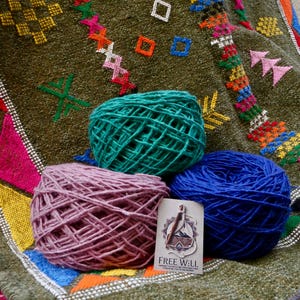 Peut inclure: Trois pelotes de laine, turquoise, violette et bleue, reposent sur un textile à motifs. Le textile est vert olive avec des motifs géométriques colorés. Une petite carte avec le texte "FREE WILL" est également présente.