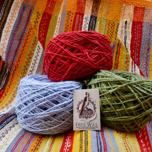 Puede incluir: Tres ovillos de lana en rojo, azul y verde sobre un textil a rayas colorido. Una pequeña tarjeta con el texto "FREE WILL" se coloca delante de la lana. La lana parece estar hilada a mano, adecuada para proyectos de punto o ganchillo.