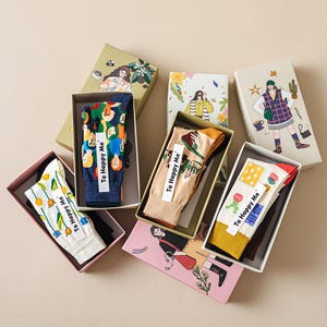 May include: A selection of patterned socks presented in illustrated gift boxes. The socks showcase diverse designs, such as floral and geometric patterns, and illustrations. Each box is adorned with a unique image, and the socks have a tag that says "To Happy Me."