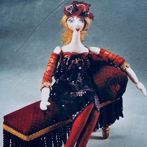 May include: A handmade doll with blonde hair and a red floral hat, seated on a burgundy bench with gold trim. The doll wears a dark dress, red stockings, and red heels with floral accents. The doll's arms are adorned with gold bracelets.