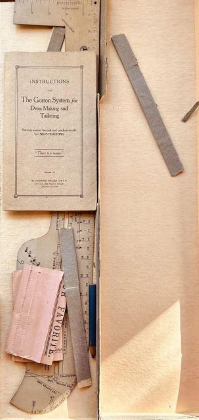 May include: A vintage Gorton System for Dress Making and Tailoring instruction booklet, a cardboard tailor's square, a curved pattern drafting tool, a small blue pencil, and a pink paper strip labeled FAVORITE, all contained within a cardboard box.