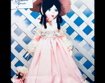 Puede incluir: Muñeca hecha a mano con un vestido rosa, un sombrero a juego y cabello negro. La muñeca tiene un cuello y puños de encaje y sostiene un pequeño bolso. El texto "Soft Porcelain Creations Janet" es visible.