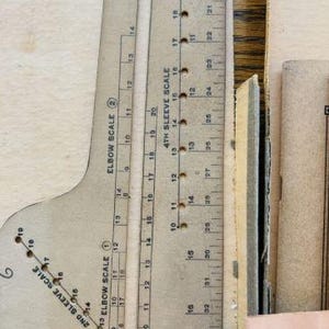 May include: A beige cardboard sewing pattern drafting tool featuring printed scales labeled Elbow Scale 2, 2nd Sleeve Scale, and 4th Sleeve Scale. The tool includes numerical markings and small circular holes for precise measurements in inches.