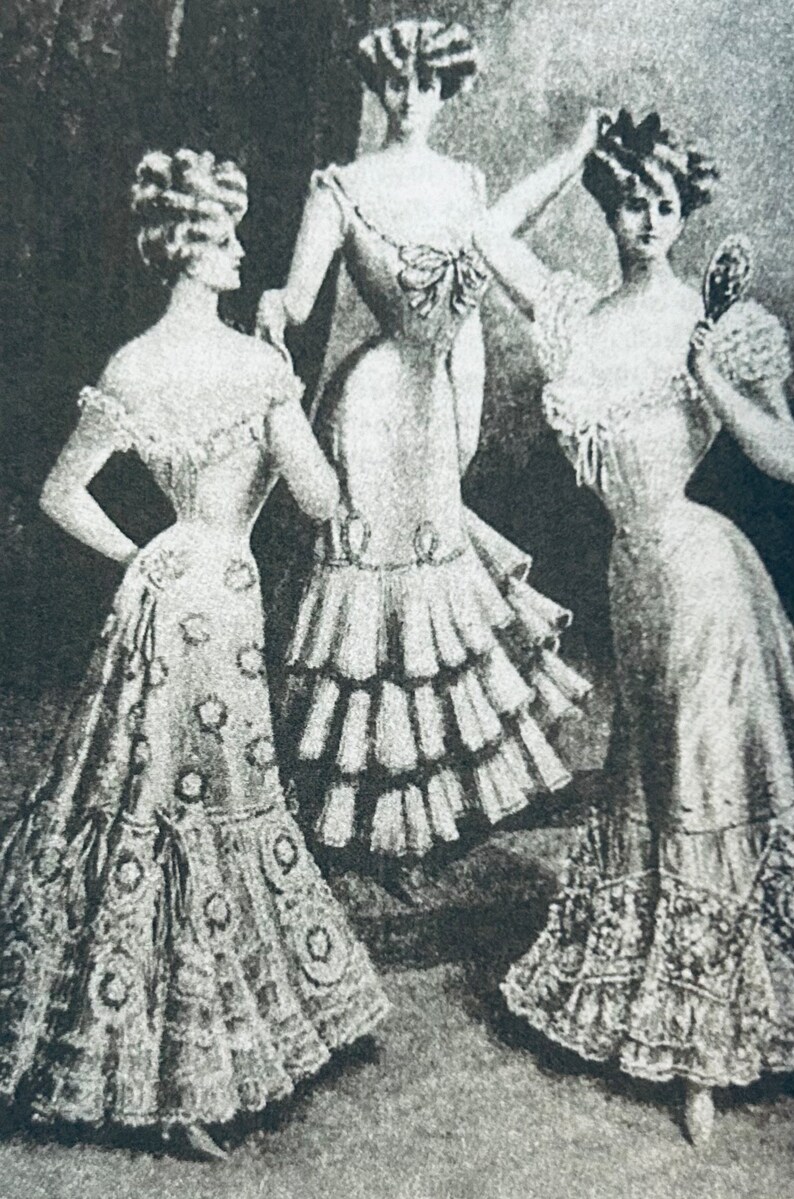 May include: Three women in vintage dresses with ruffled skirts and elaborate hairstyles. The women are standing in a studio setting and are looking at the camera.